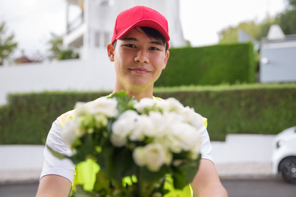 A delivery person wearing a bright yellow vest and a red cap is smiling while holding out a bouquet of fresh white flowers towards the camera. The bouquet includes a variety of blooms with delicate pe