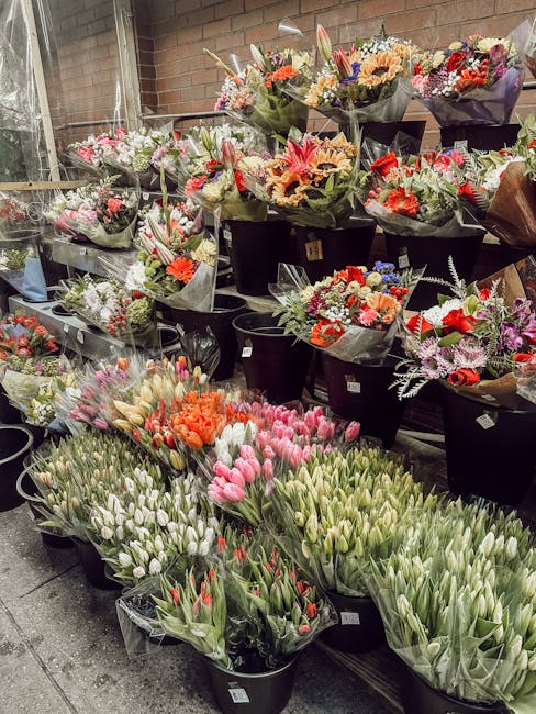 A display of fresh flower bouquets arranged in black buckets and wrapped in decorative paper, featuring a variety of flowers including tulips in shades of pink, white, and red, as well as carnations a