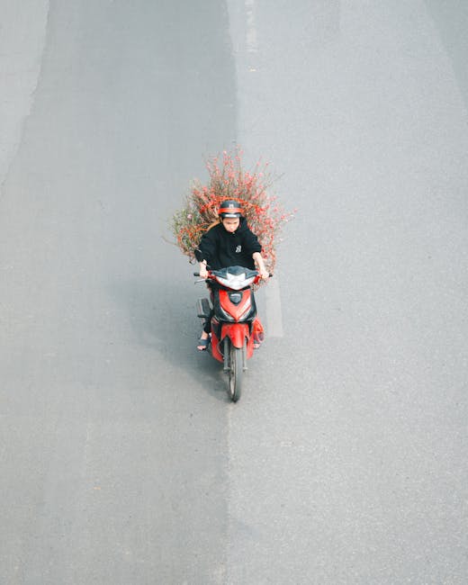 A person wearing a black jacket and helmet rides a red scooter along a grey urban street, carrying a large bouquet of fresh flowers arranged with pink and red blossoms, greenery, and delicate sprigs o