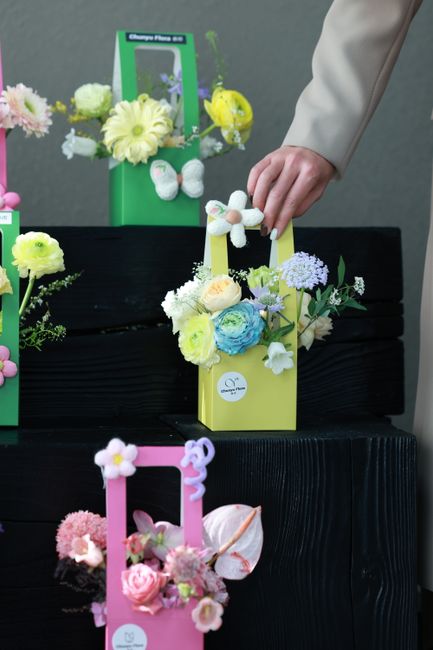 A display of four floral arrangements in colorful, rectangular cardboard boxes—green, yellow, pink, and purple—each containing a variety of fresh flowers including white roses, blue and purple hyd