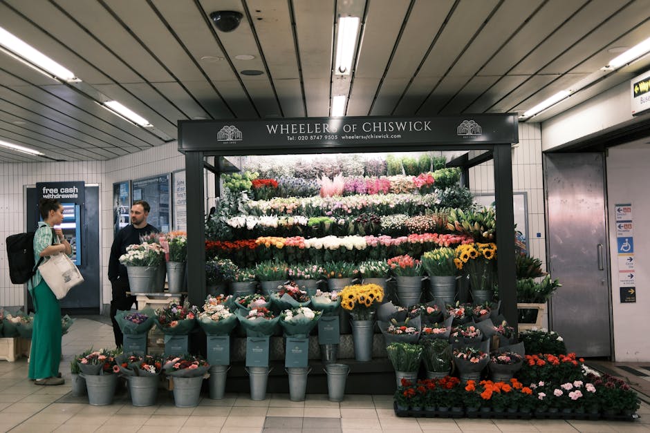 A vibrant floral display at Wheeler of Chiswick, situated in an underground station, showcasing a variety of fresh flowers arranged in buckets and pots. The arrangement includes bright yellow sunflowe