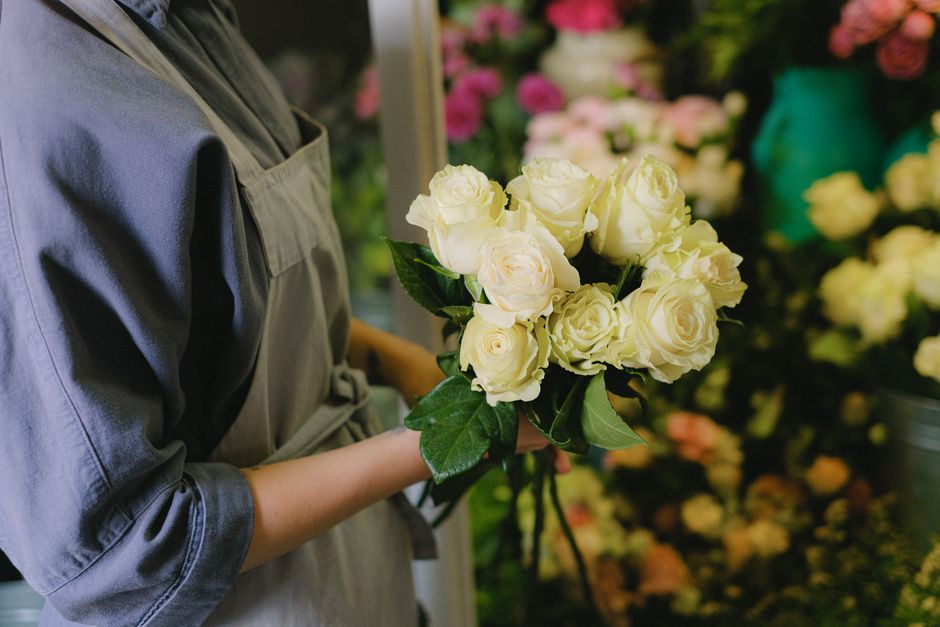 A person dressed in a grey apron holding a bouquet of fresh, cream-colored roses with lush green leaves in both hands, standing in a floral shop. The bouquet features tightly clustered roses with slig